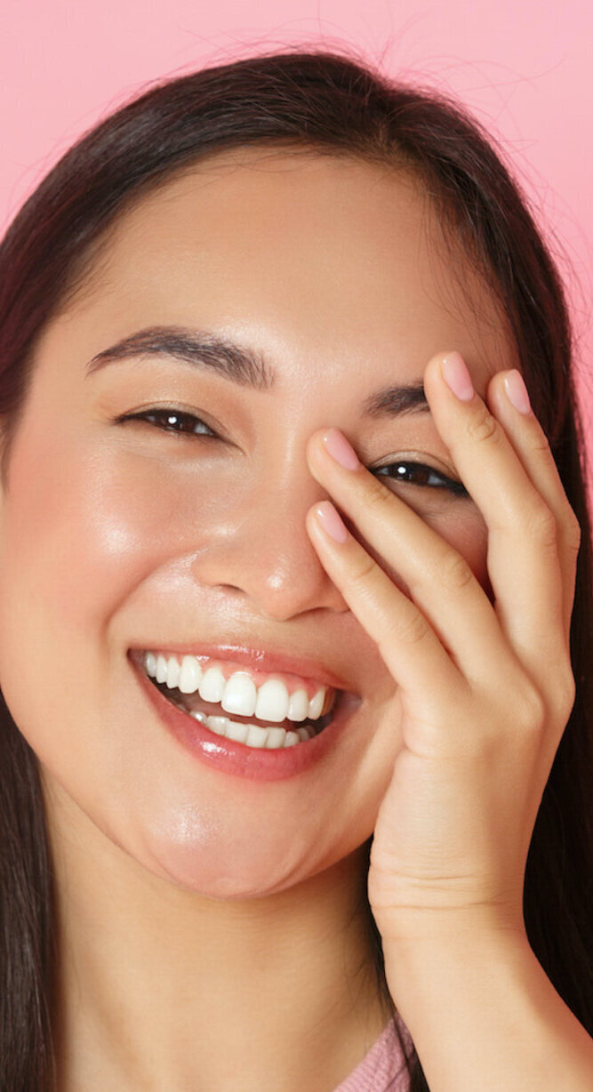 Beauty, fashion and lifestyle concept. Close-up of carefree beautiful asian girl touching face and laughing happily with closed eyes, standing over pink background joyful, skincare product promo